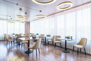 Uma sala de conferências com várias mesas e cadeiras, grandes janelas com cortinas e luminárias de teto circulares.