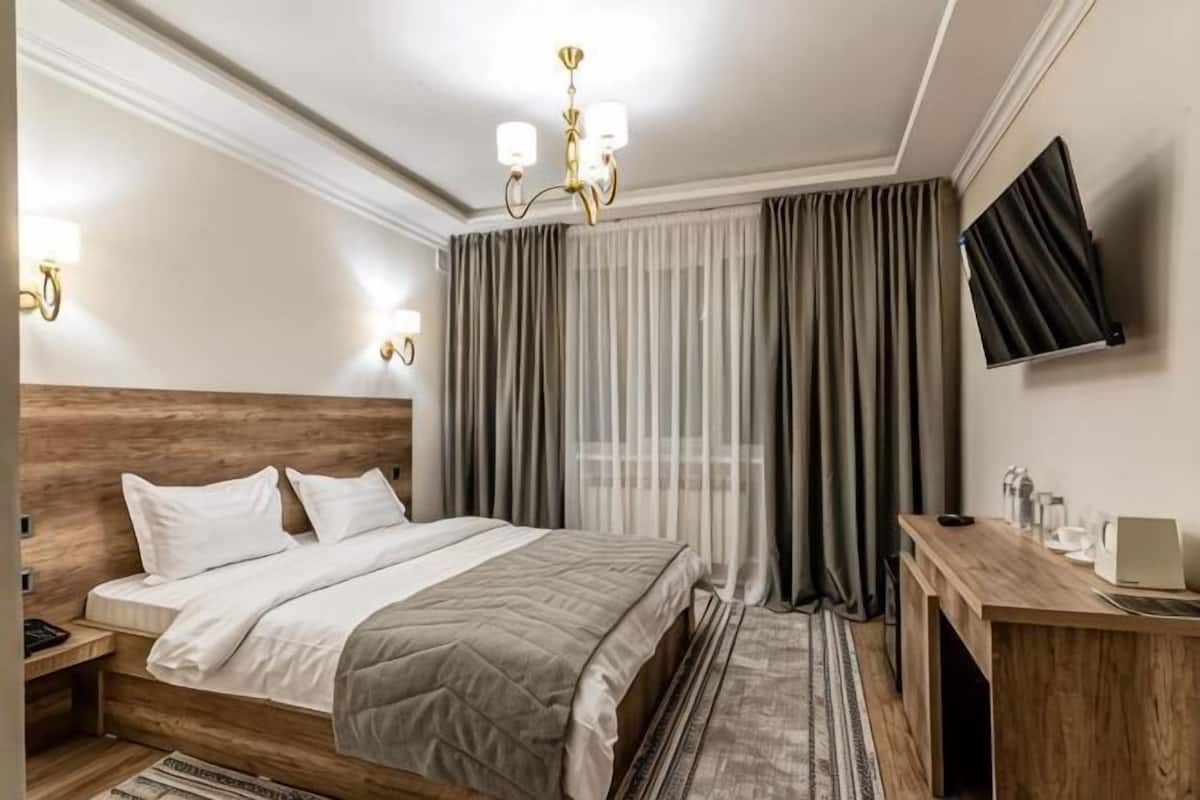 Habitación de hotel con cabecera de madera, cama con ropa blanca, escritorio con silla, televisor montado en la pared y ventana con cortinas.