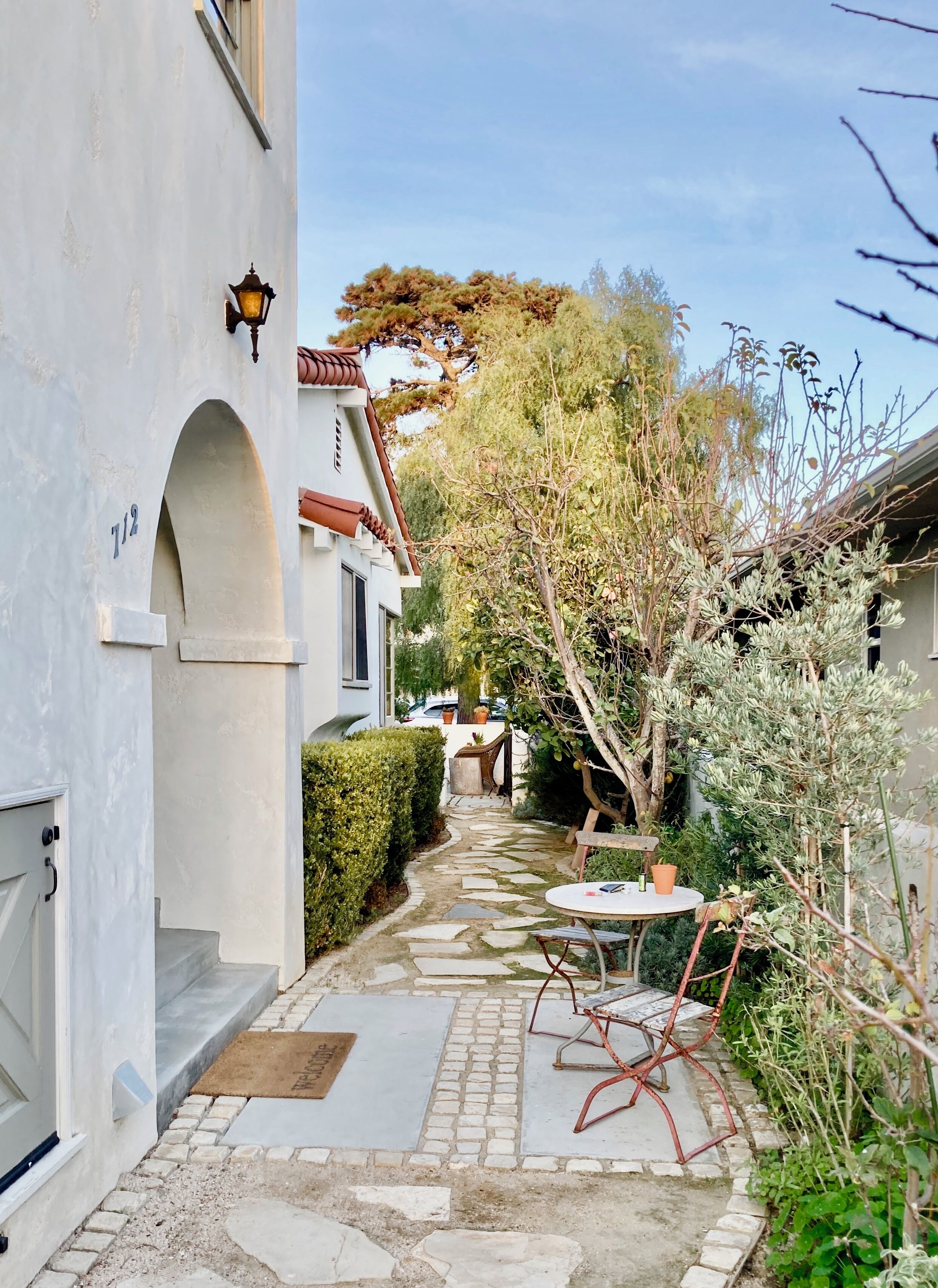 A narrow stone pathway leading to a white building with a number '712' sign. There is a small round table with chairs and a potted plant nearby.