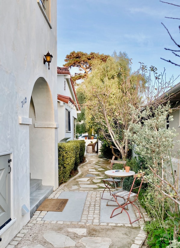 A narrow stone pathway leading to a white building with a number '712' sign. There is a small round table with chairs and a potted plant nearby.