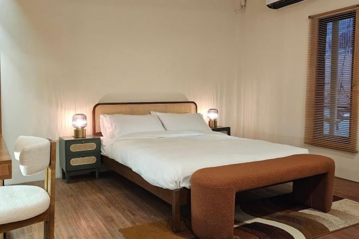 Un dormitorio con cama, mesitas de noche, un banco, una silla y una ventana con persianas.