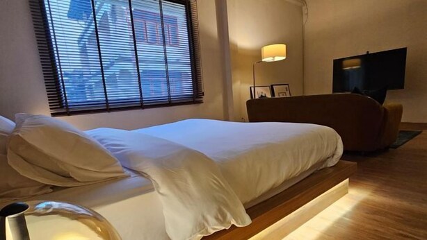 A modern hotel room with a bed, bedside lamp, and a large window with blinds. - Yaowarat Stay (Bangkok)