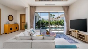 A modern living room with a large sofa, a flat-screen TV, and a view of a pool and palm trees. - Two Flamingo Villa 2 (Phuket)