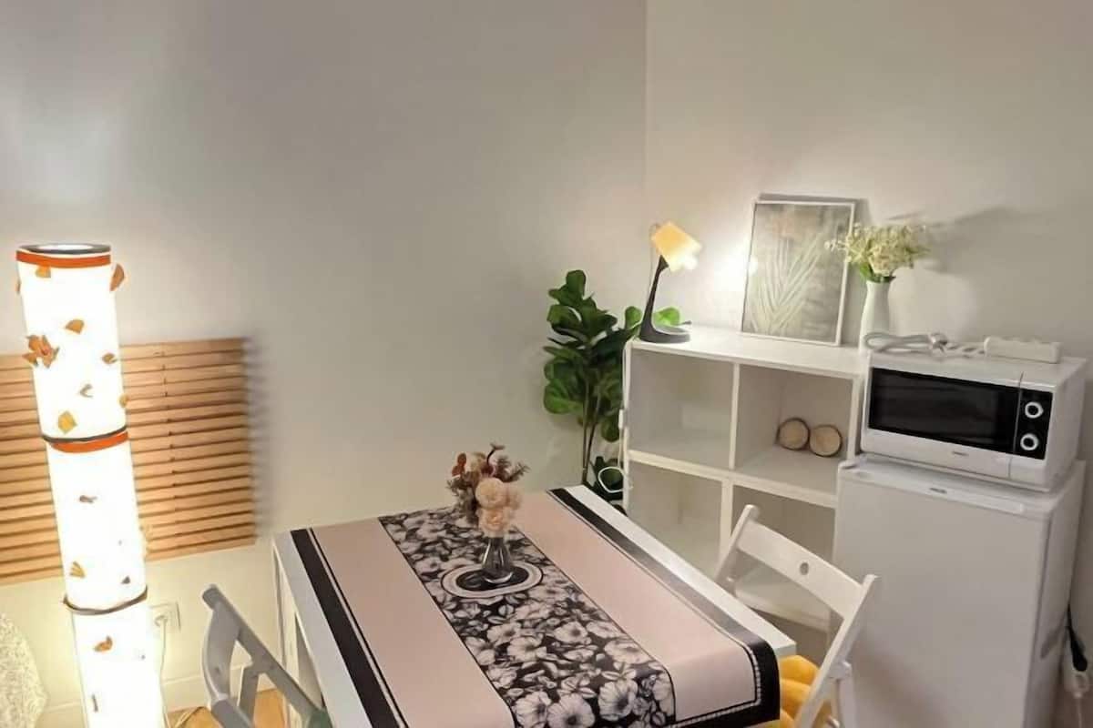 Una cocina compacta con microondas, un refrigerador pequeño, una mesa de comedor con mantel floreado y una lámpara de pie alta.