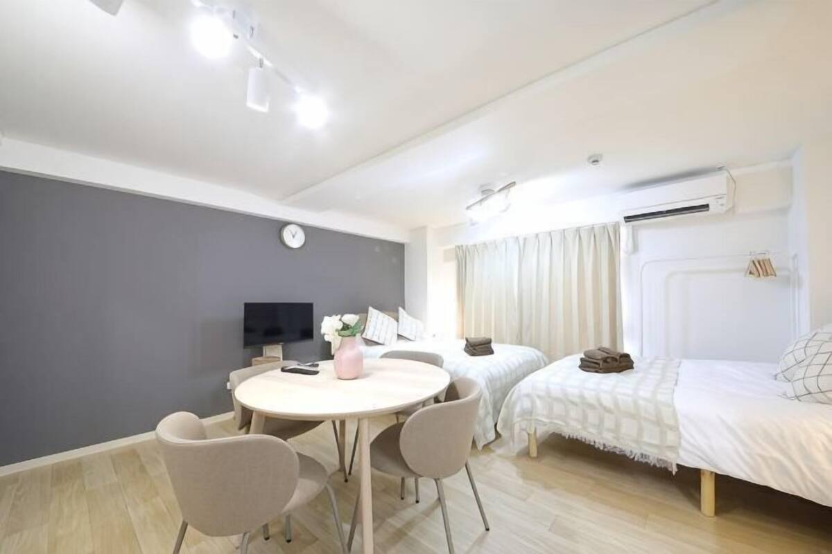 A modern hotel room with a round dining table, two single beds, a TV, and a clock on the wall.