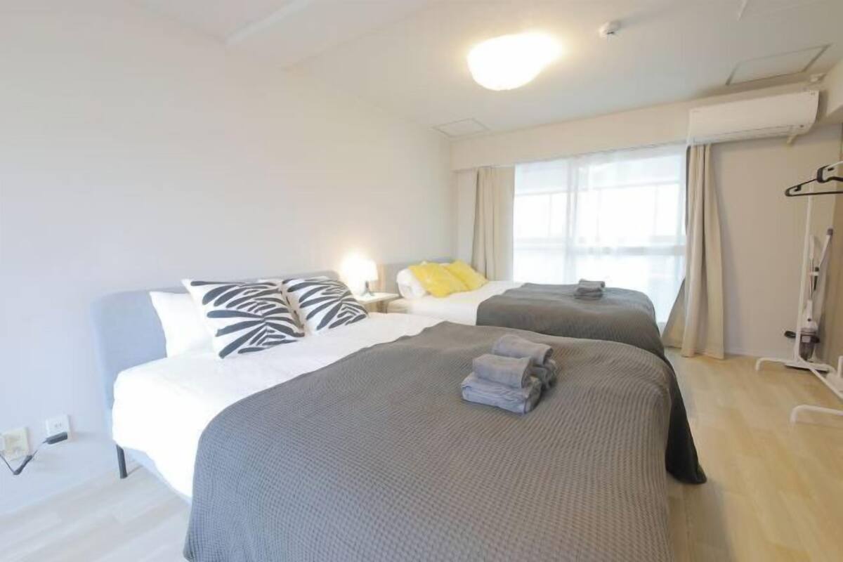 A hotel room with two beds, a window with curtains, a bedside table with a lamp, and a wall-mounted air conditioner.
