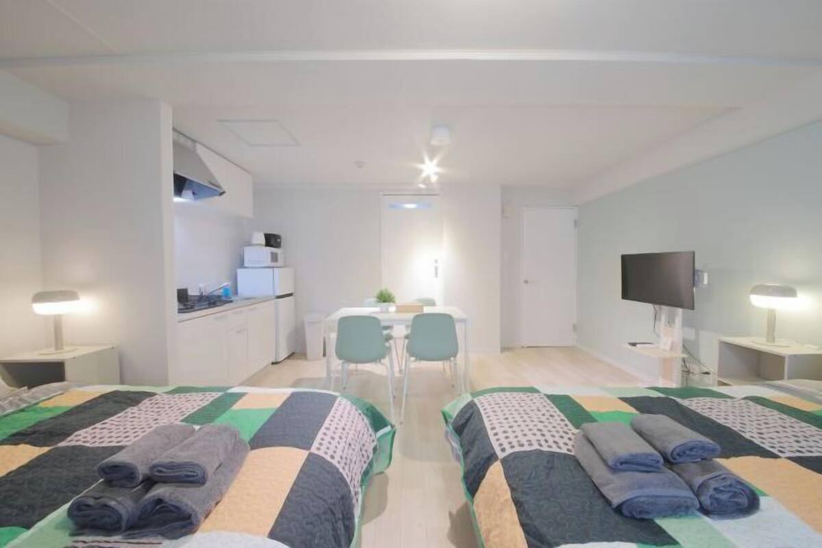 A hotel room with two beds, a desk, a TV, and a kitchenette.