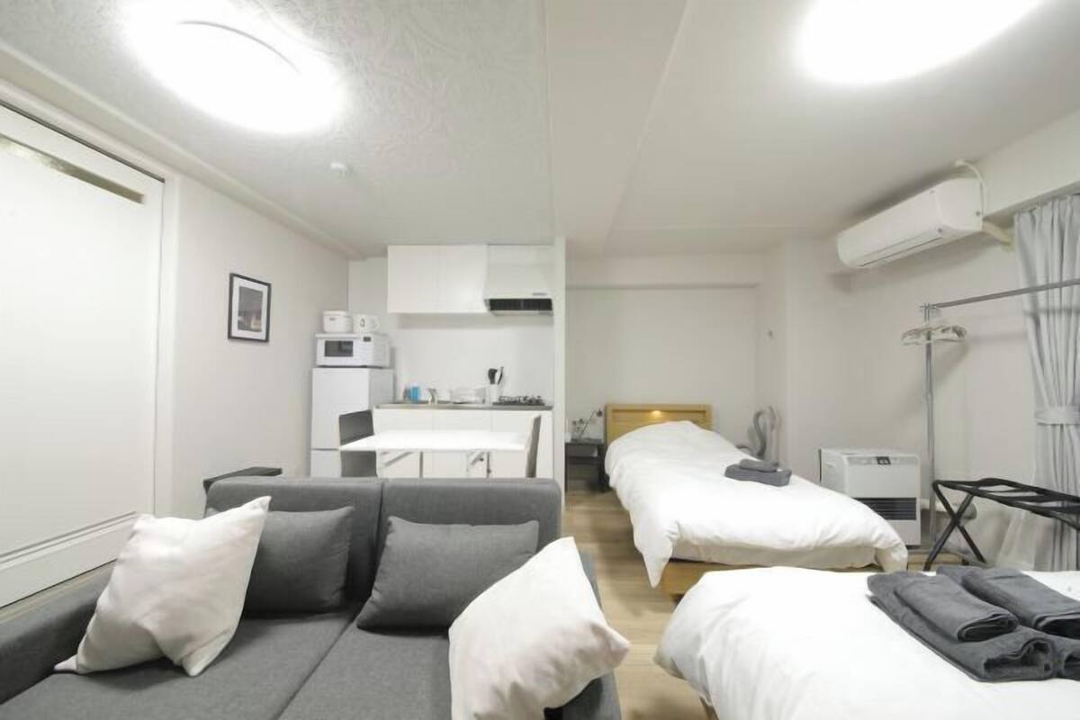 A compact hotel room with a grey sofa, two single beds, a small kitchenette, and a ceiling fan.