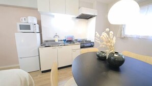 A compact kitchen with a microwave, stove, and sink. There is a dining table with chairs and a vase with dried flowers. - Mansion Cattleya. (Sapporo)