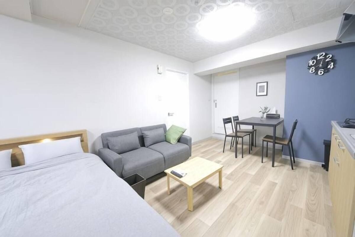 A room with a bed, a gray sofa, a small table, a dining area with chairs, and a kitchen area.