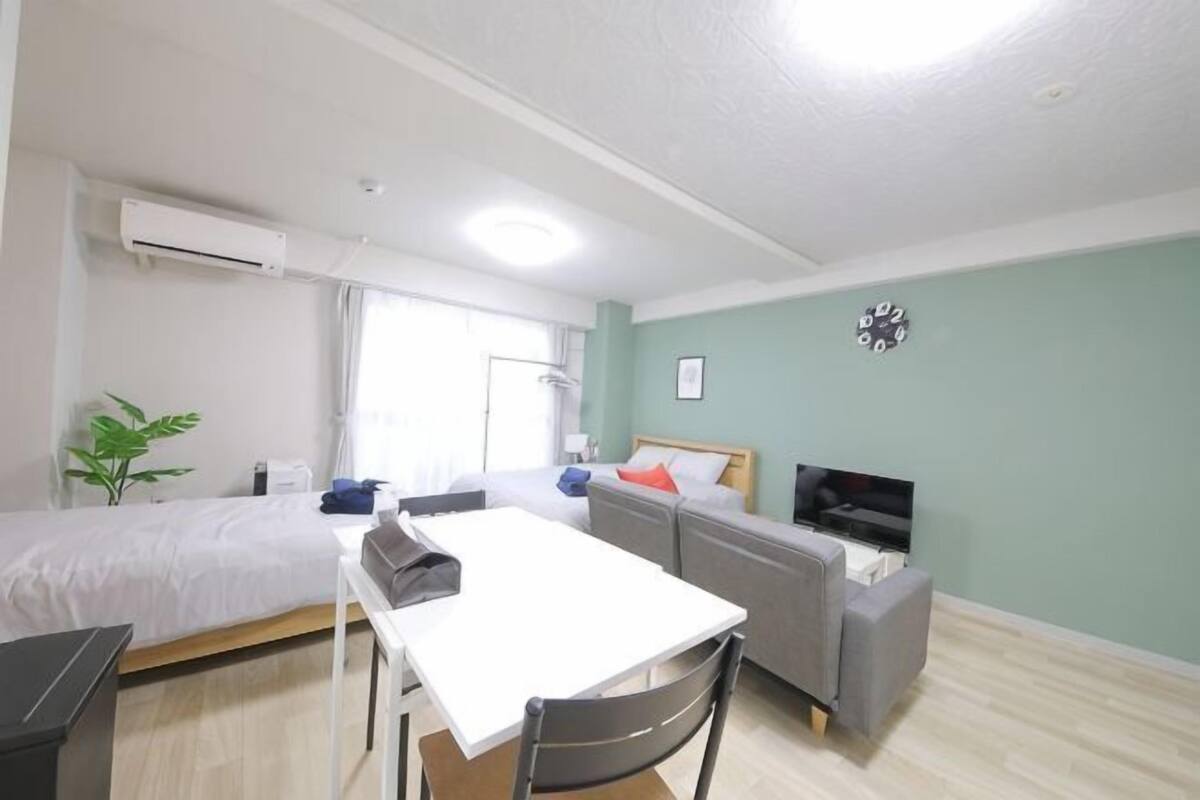 A room with two beds, a table, a chair, a TV, and a plant.