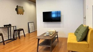 A modern living room with a flat-screen TV mounted on the wall, a wooden coffee table, a yellow sofa, and a small side table with a lamp. - ASOBO OSAKA Kuromon 3rd (Osaka)