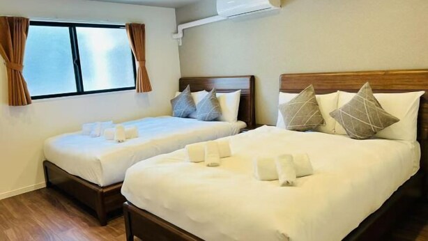 A hotel room with two beds, a window with curtains, an air conditioning unit, and wooden flooring. - ASOBO OSAKA Kuromon 3rd (Osaka)