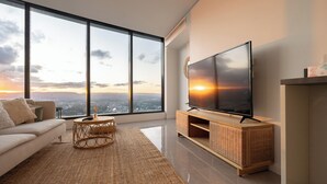 A modern living room with a large TV, a sofa, a round coffee table, and a view of the city at sunset. - Sunset Boulevard - Sky High Apt on Ocean Meirton (Surfers Paradise)