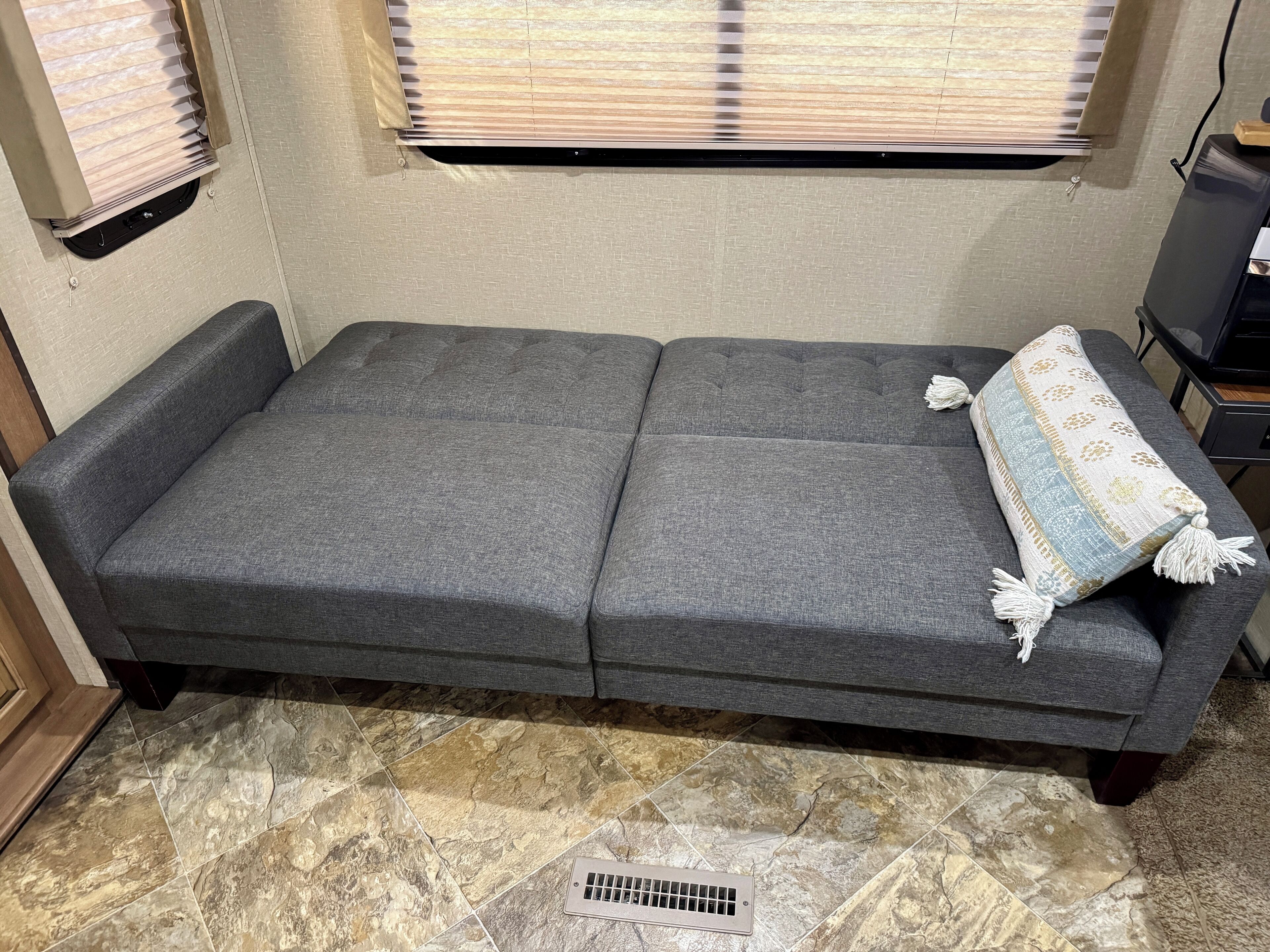 A grey sofa with a patterned throw blanket, a tiled floor, and a window with blinds.