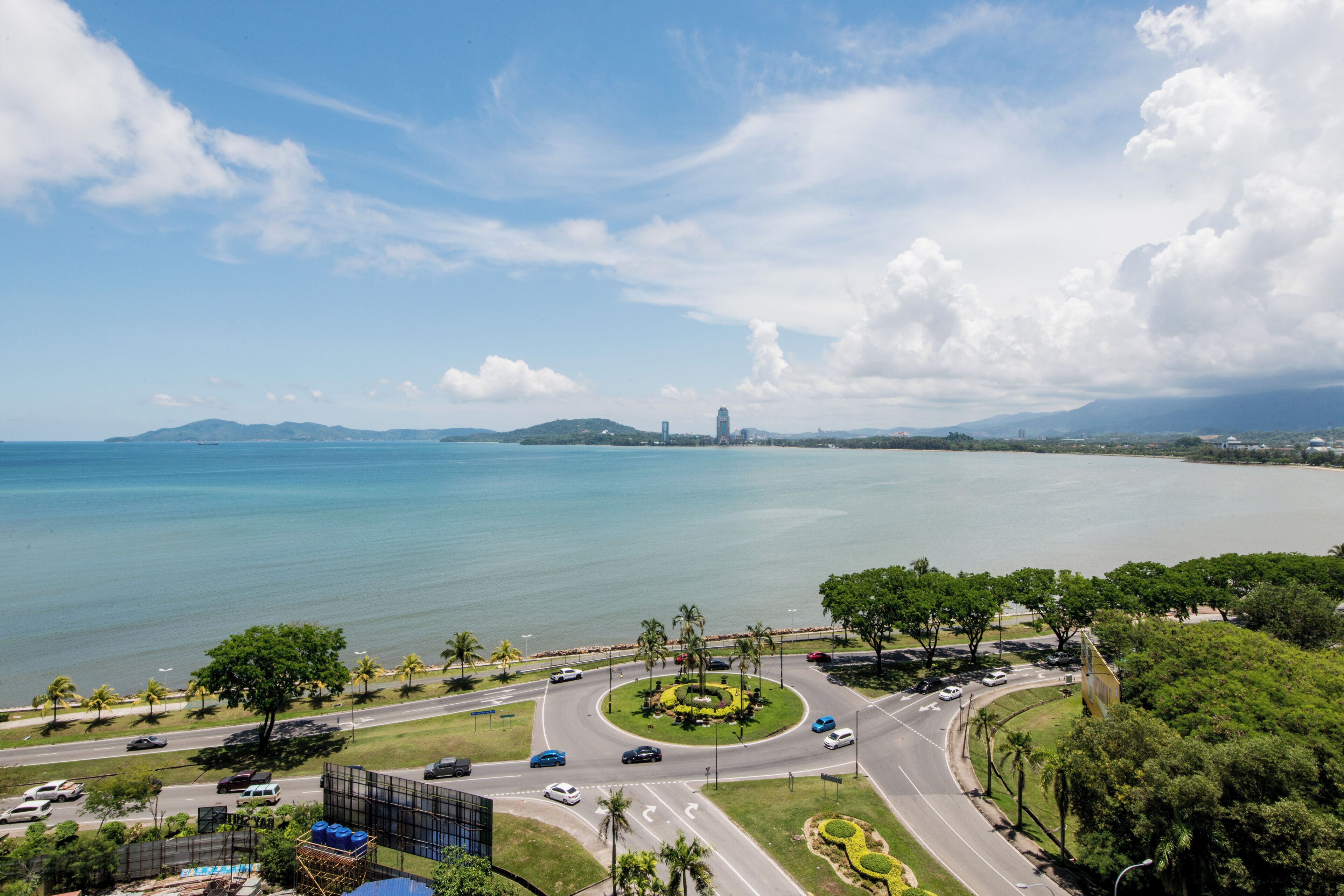 A coastal road with a roundabout, palm trees, and a view of the sea and distant mountains.