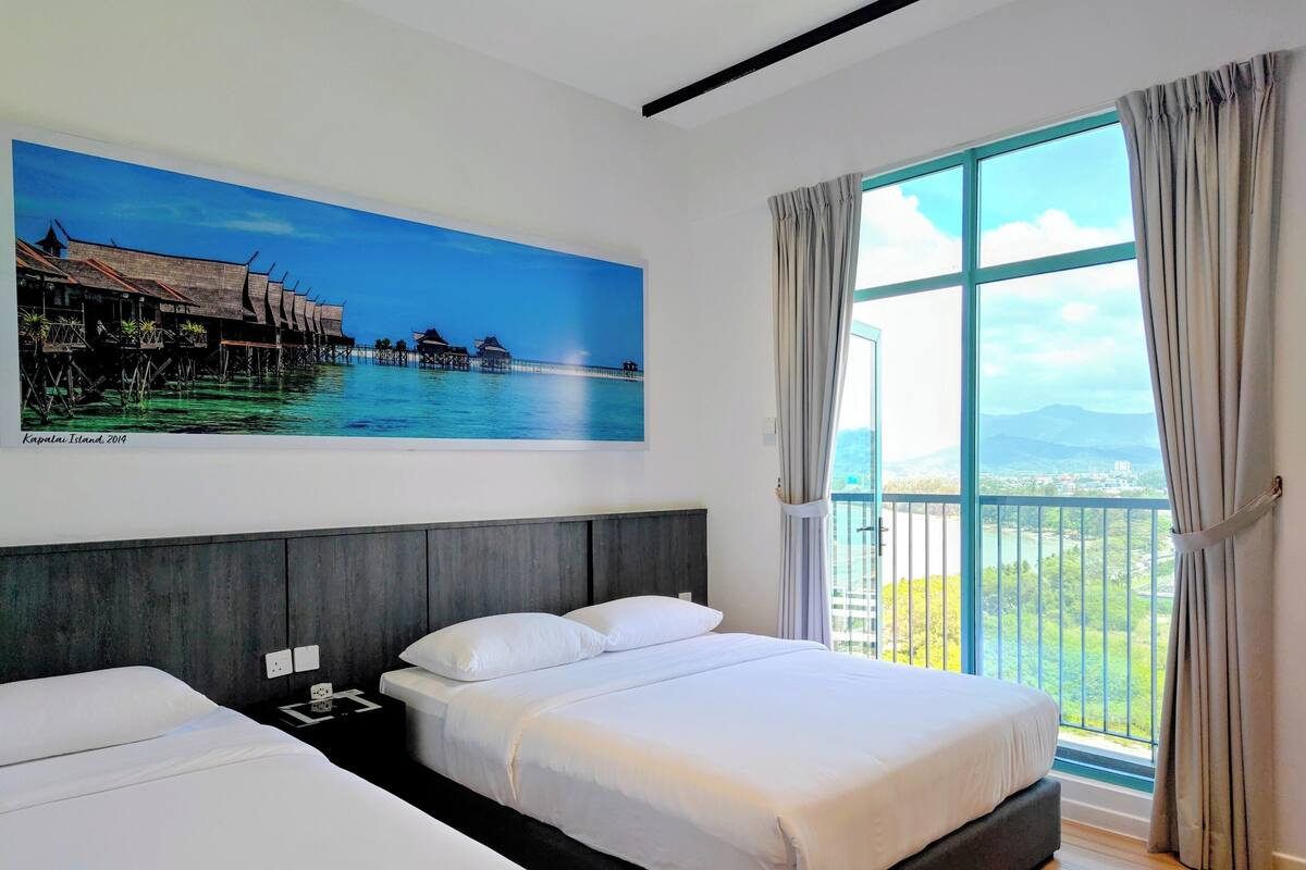 A hotel room with two beds, a large window with curtains, a painting of a coastal scene, and a balcony with a view.