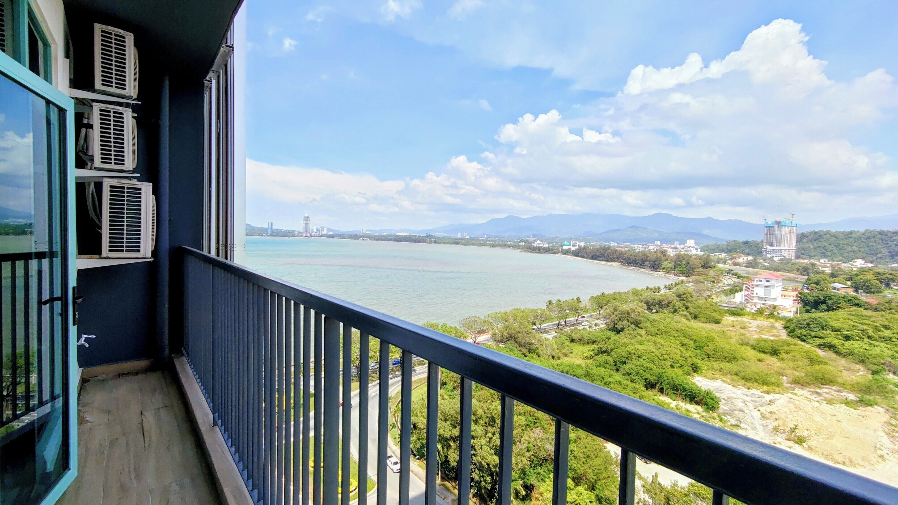 A balcony with air conditioning units, overlooking a body of water with a distant cityscape and mountains.