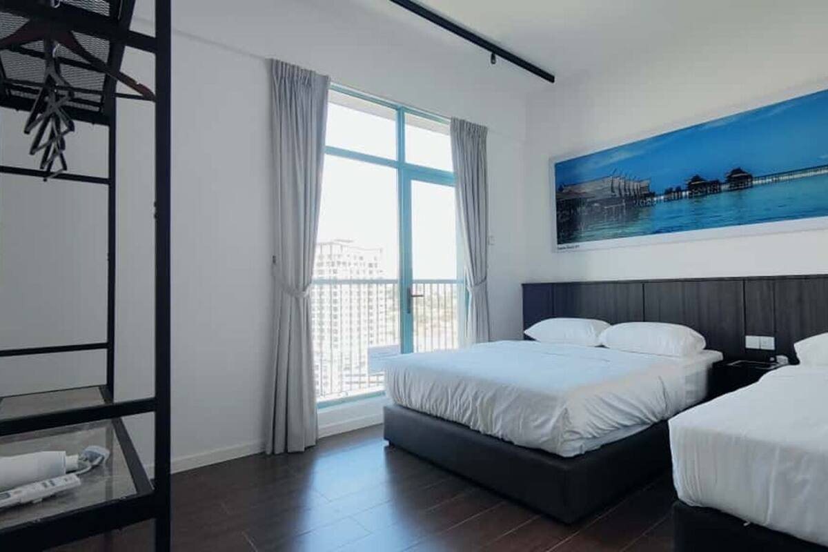 A hotel room with two beds, a large window with curtains, a painting of a coastal scene, and a metal rack with towels.
