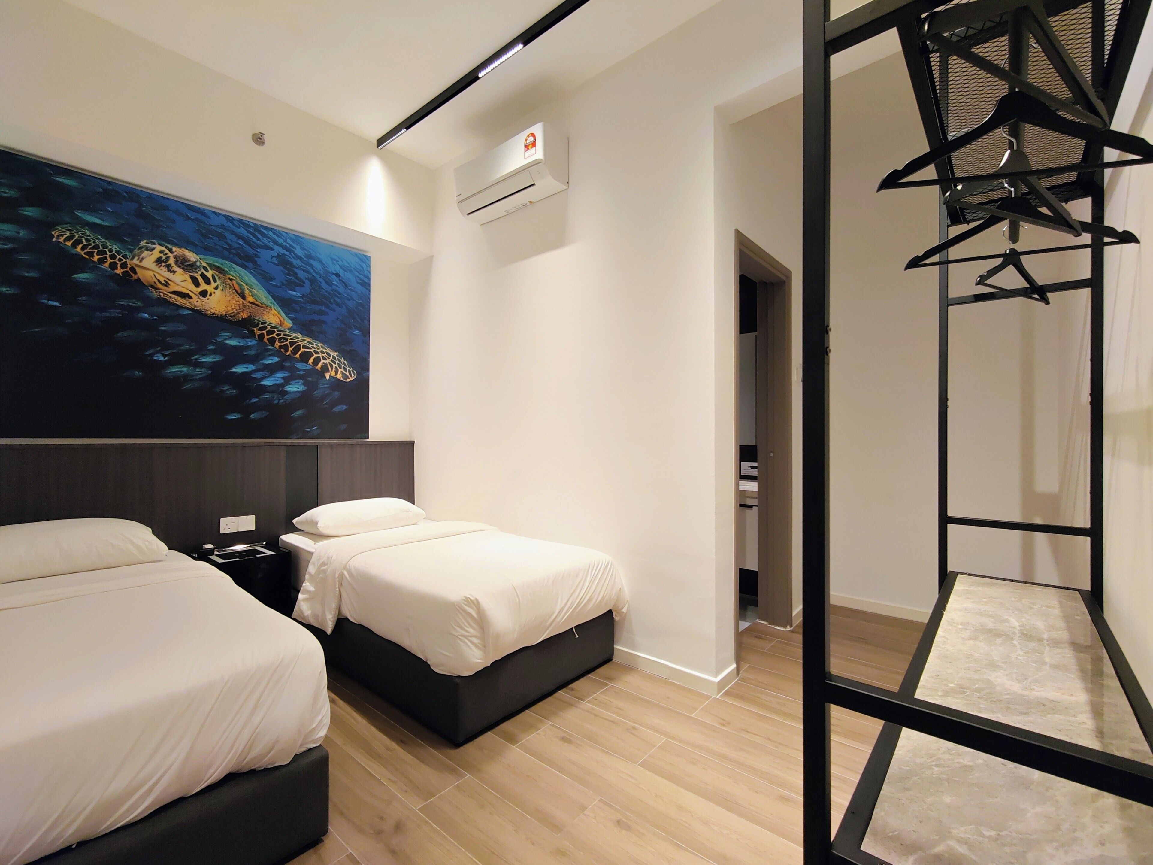 Quarto de hotel com duas camas, um grande mural na parede de uma tartaruga marinha, um aparelho de ar condicionado e um suporte de metal preto.