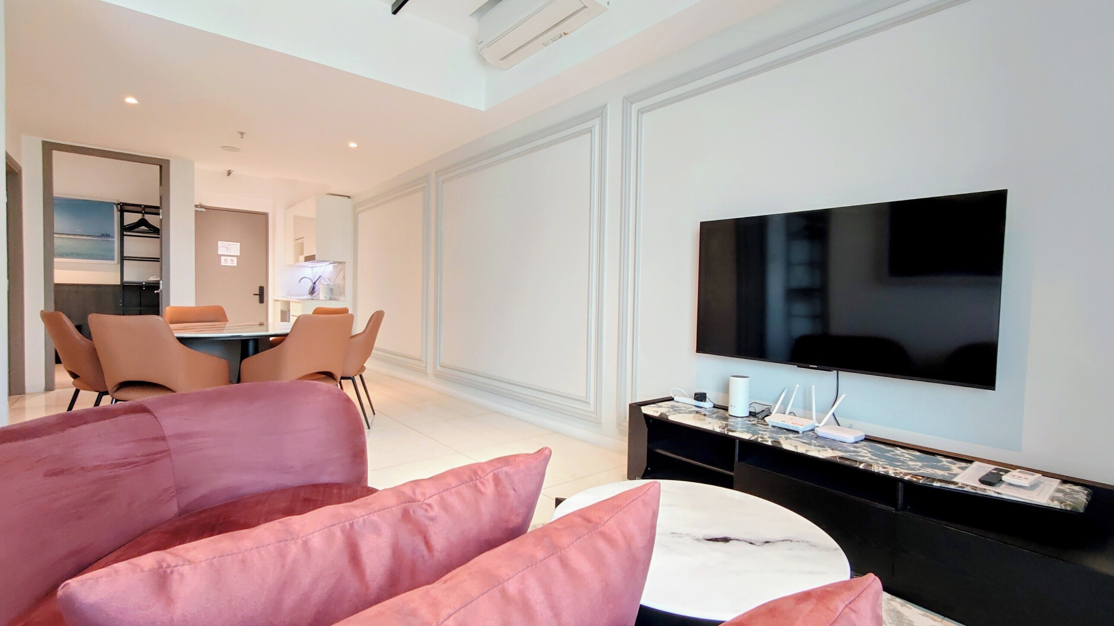 A modern hotel room with a flat-screen TV, a dining area, and a seating area with a pink sofa.
