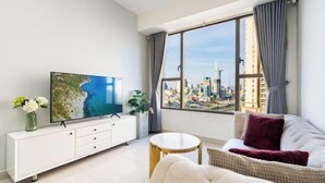 A modern living room with a large flat-screen TV, a white cabinet, a round coffee table, a sofa, and a view of a cityscape through a large window. - MIN' Apartment - Rivergate Residence Downtown BUI VIEN - FREE POOL (Ho Chi Minh City)