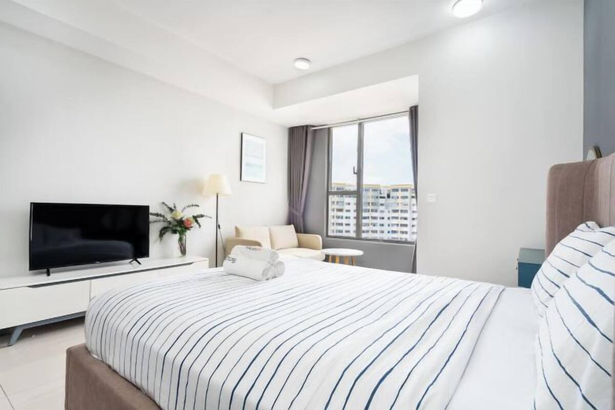 Ein modernes Hotelzimmer mit einem großen Bett, einem Flachbildfernseher, einem Nachttisch mit Lampe und Blick auf die Stadt durch das Fenster.