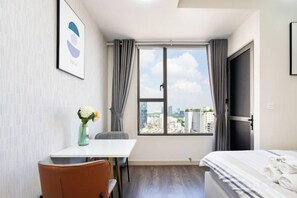 A hotel room with a bed, a desk, a chair, a window with curtains, and a city view. - MIN' Apartment - Rivergate Residence Downtown BUI VIEN - FREE POOL (Ho Chi Minh City)