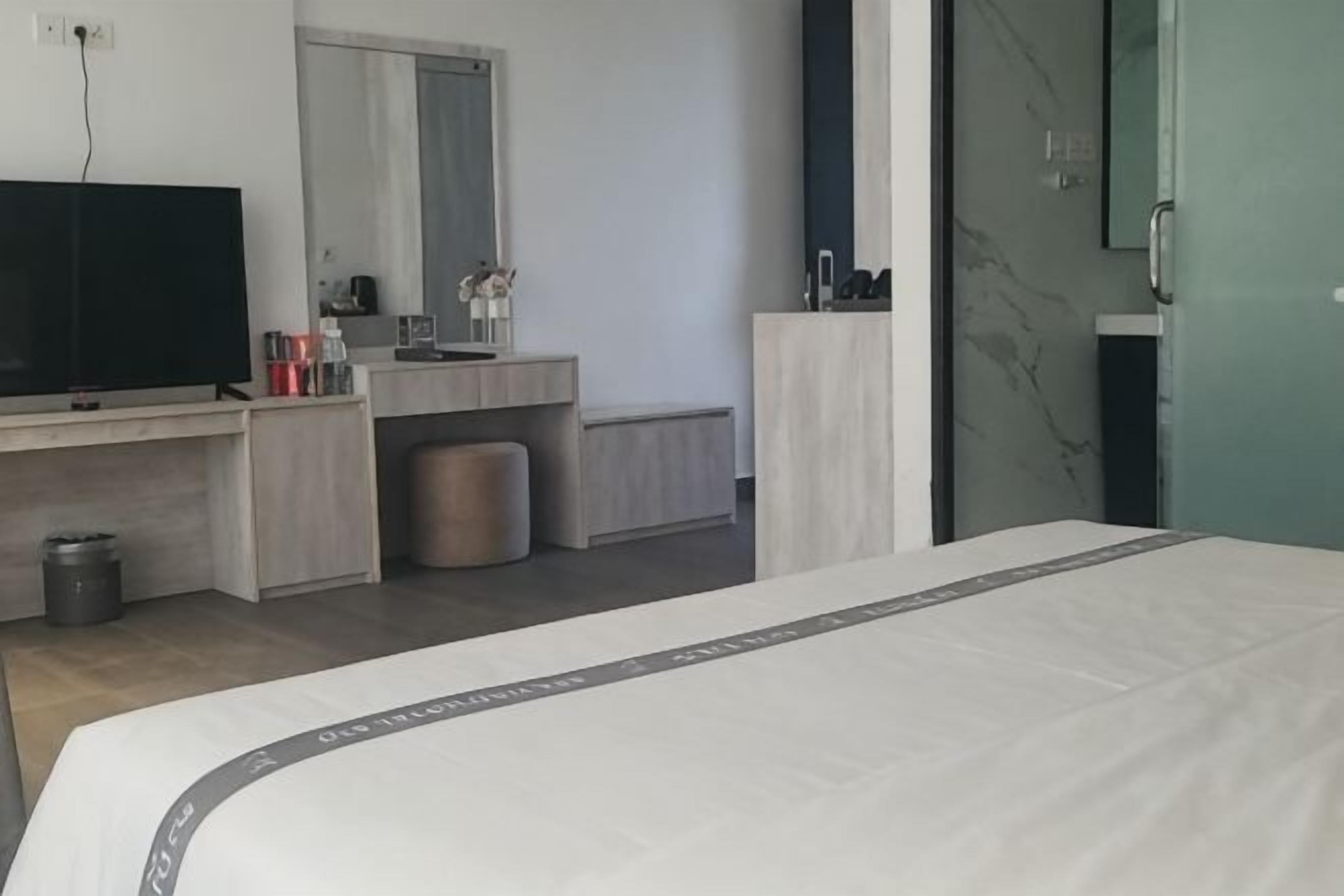 Ein modernes Hotelzimmer mit einem Bett, einem Fernseher, einem Schminktisch mit Spiegel, einer kleinen Mülltonne und einer Tür mit Griff.
