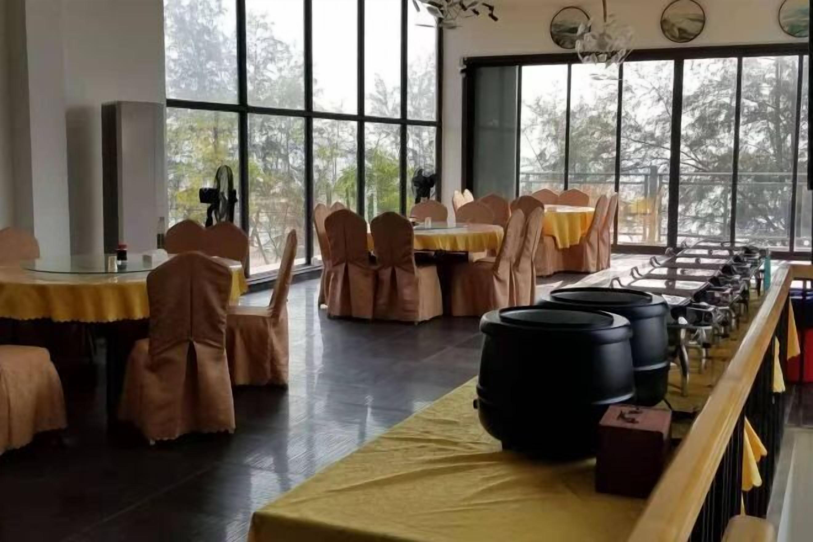 A restaurant with round tables covered with yellow tablecloths and brown chairs. Large windows provide a view of trees outside.