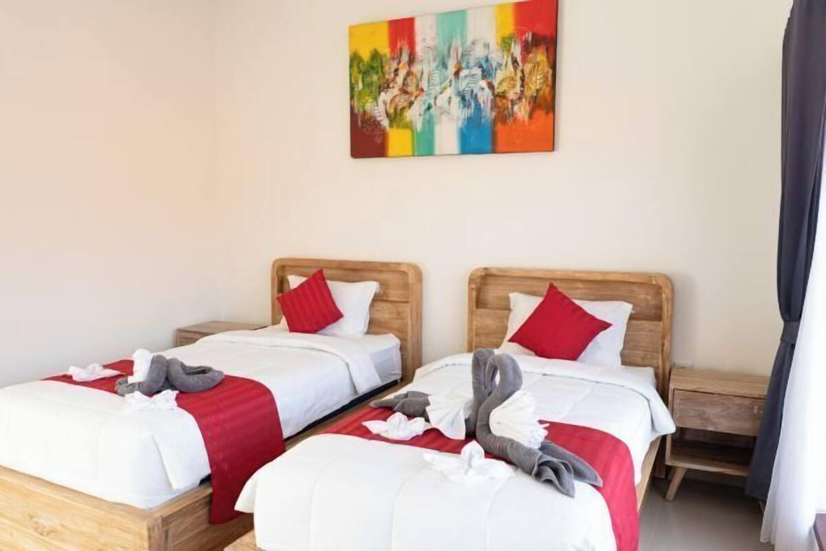Une chambre d’hôtel avec deux lits simples, des têtes de lit en bois, du linge de lit blanc, des oreillers rouges et une peinture abstraite aux couleurs vives accrochée au mur.