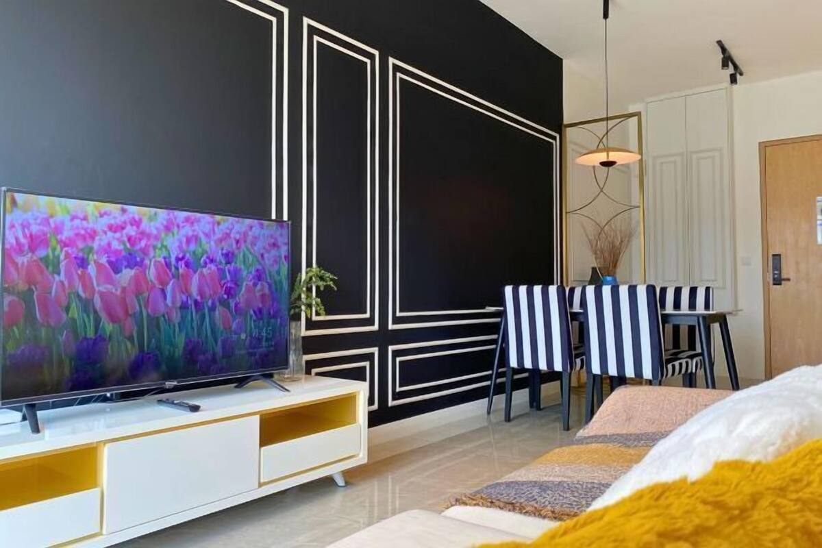 Una sala de estar moderna con un televisor de pantalla plana, una zona de comedor con sillas a rayas y un diseño de pared geométrico en blanco y negro.