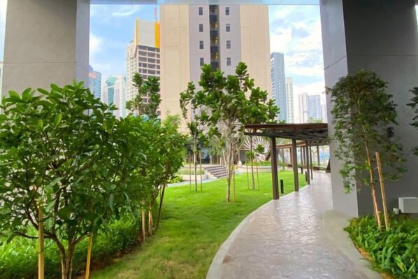 Un chemin bordé de verdure et agrémenté d’une pergola, qui mène à un bâtiment au design moderne offrant une vue sur des immeubles de grande hauteur.