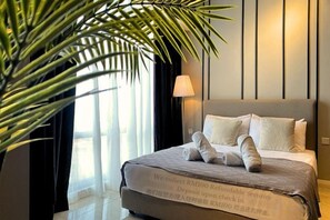 Une chambre d’hôtel avec un lit, une grande plante en pot, une lampe et une fenêtre avec des rideaux.