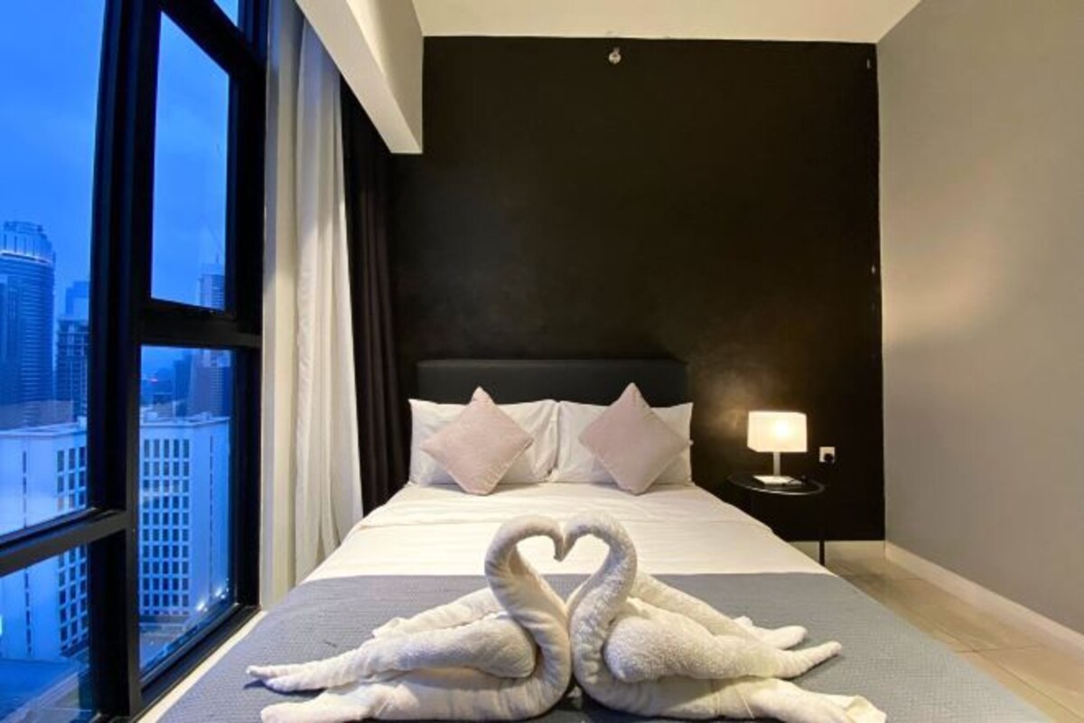 A hotel room with a large window, a bed with white linens, a nightstand with a lamp, and a heart-shaped towel arrangement on the bed.