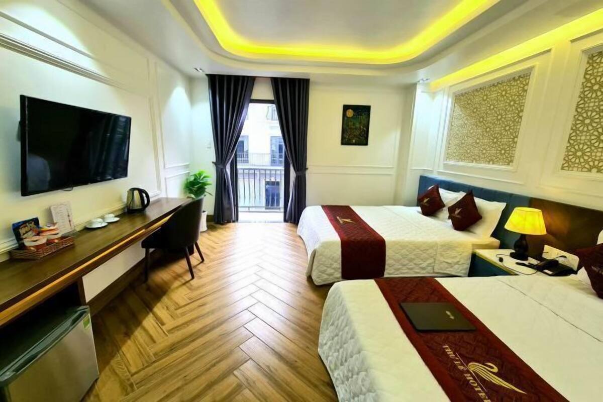 A hotel room with a flat-screen TV, a desk with a computer, two beds with red bedspreads, a nightstand with a lamp, and a view of the outside balcony.