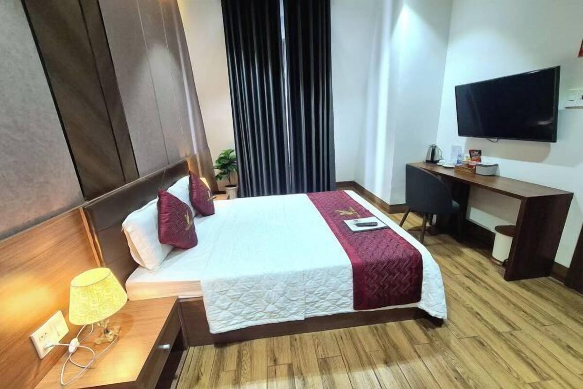 A hotel room with a bed, bedside table with a lamp, a desk with a chair, a television on the wall, and a window with curtains.