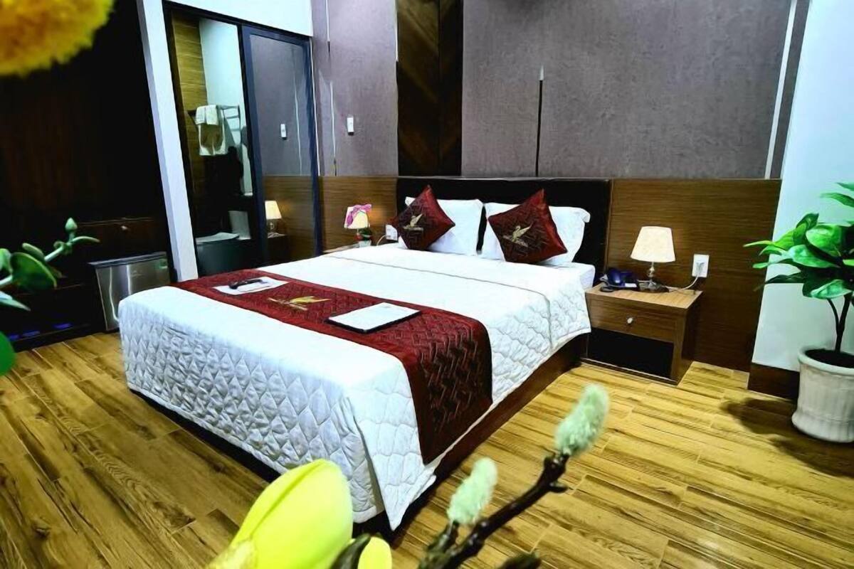 A hotel room with a large bed, wooden flooring, a nightstand with a lamp, a mirror, and a potted plant.