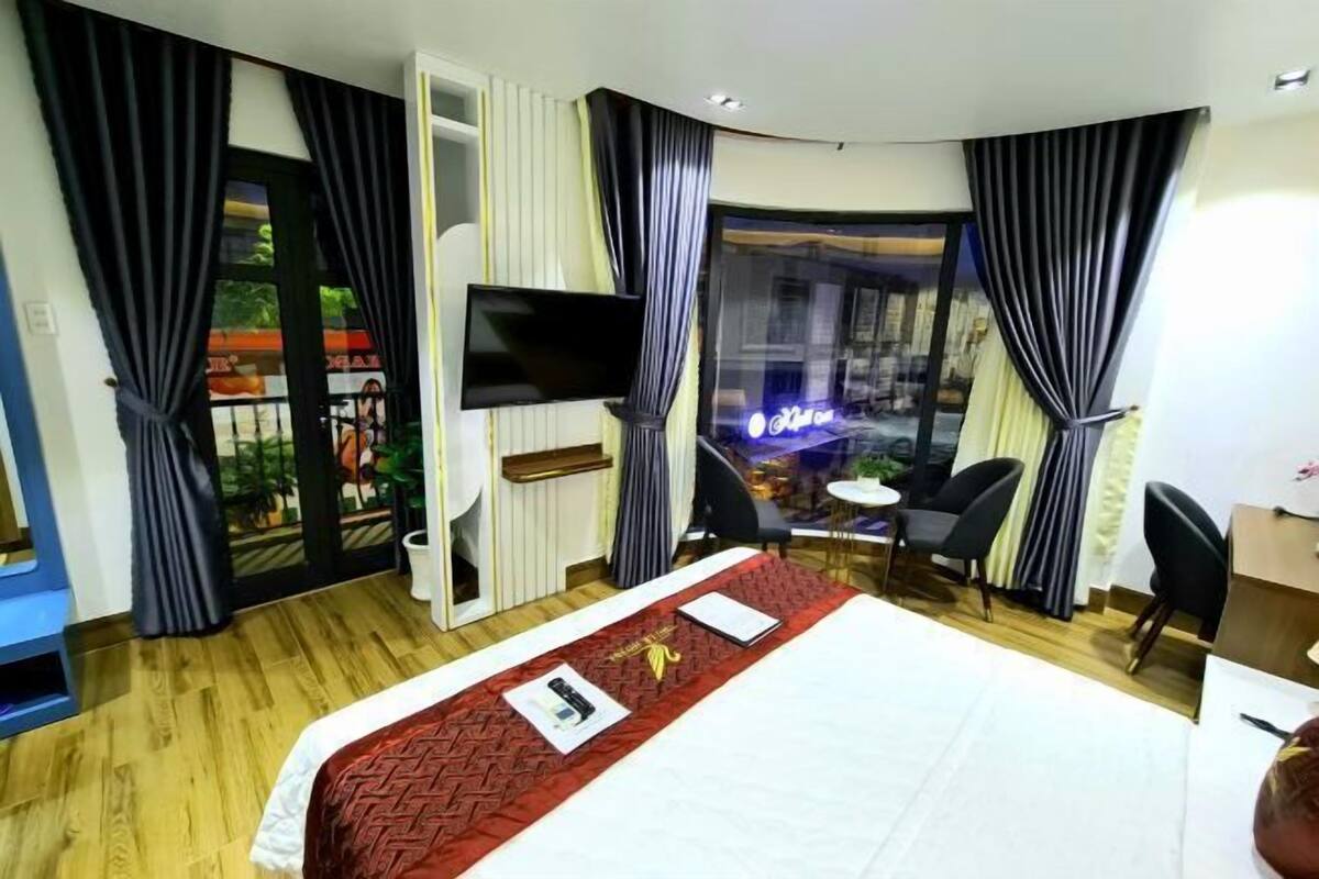 A hotel room with a large bed, a flat-screen TV mounted on the wall, a small table with chairs, and a view of a cityscape through large windows.