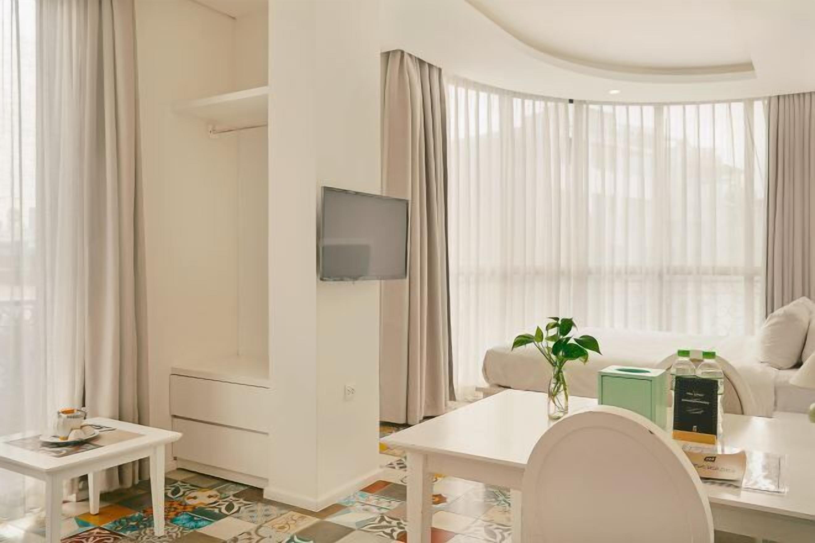 Ein modernes Hotelzimmer mit einem Wand-Fernseher, einem weißen Sofa, einem kleinen Tisch mit Blumenvase und einem Esstisch mit einer grünen Kiste und einer Flasche.