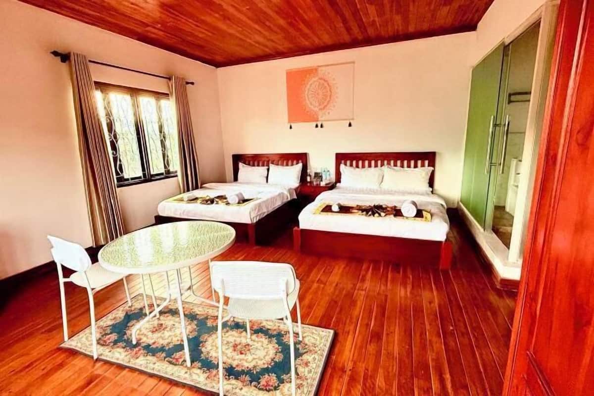 A room with two beds, a round table with chairs, a window with curtains, and a green wardrobe.