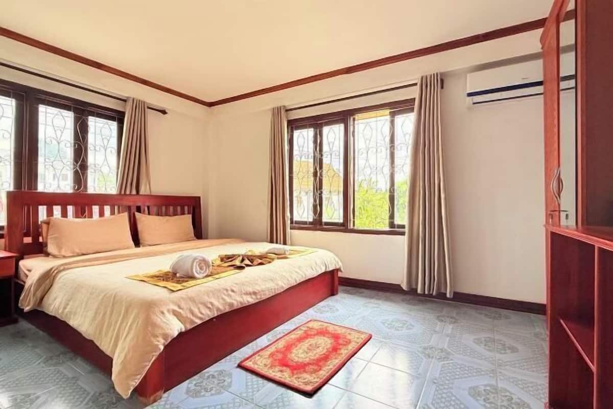 A bedroom with a wooden bed, beige bedding, a red and beige rug, and patterned floor tiles.