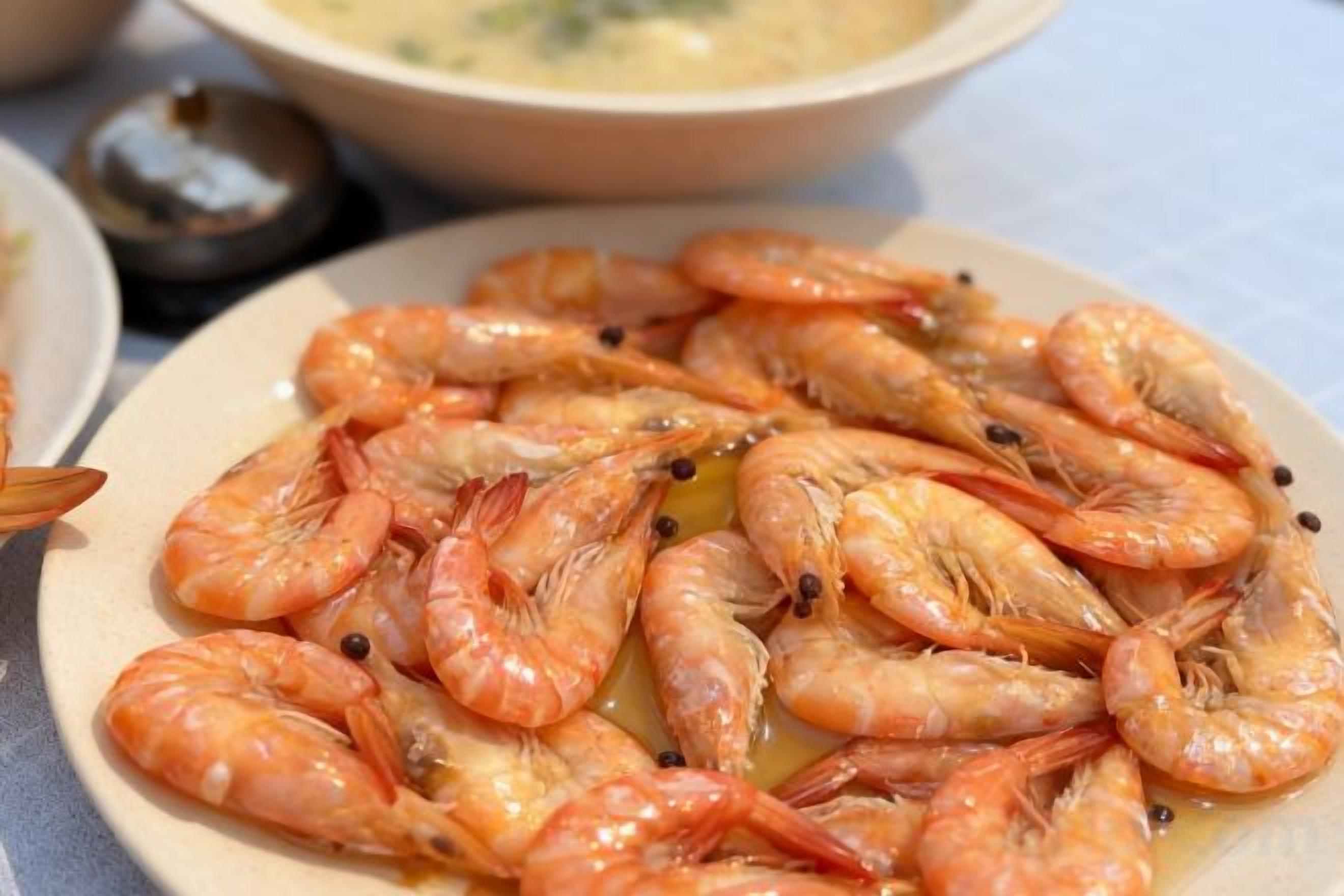 A plate of cooked shrimp with a sauce, accompanied by a bowl of soup and a small container of condiments.