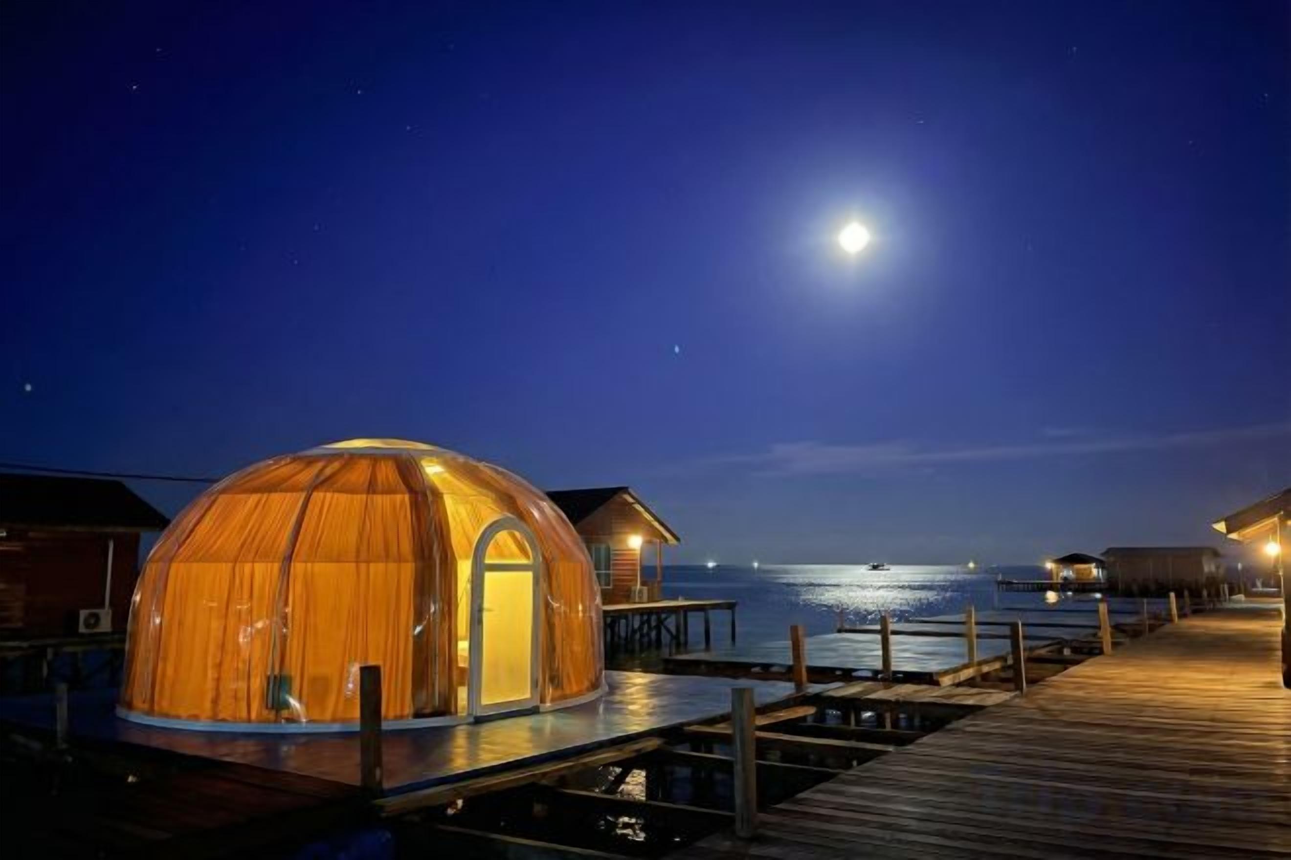 A dome-shaped structure with a door, connected to a wooden dock, overlooking a calm body of water under a night sky with a visible moon.