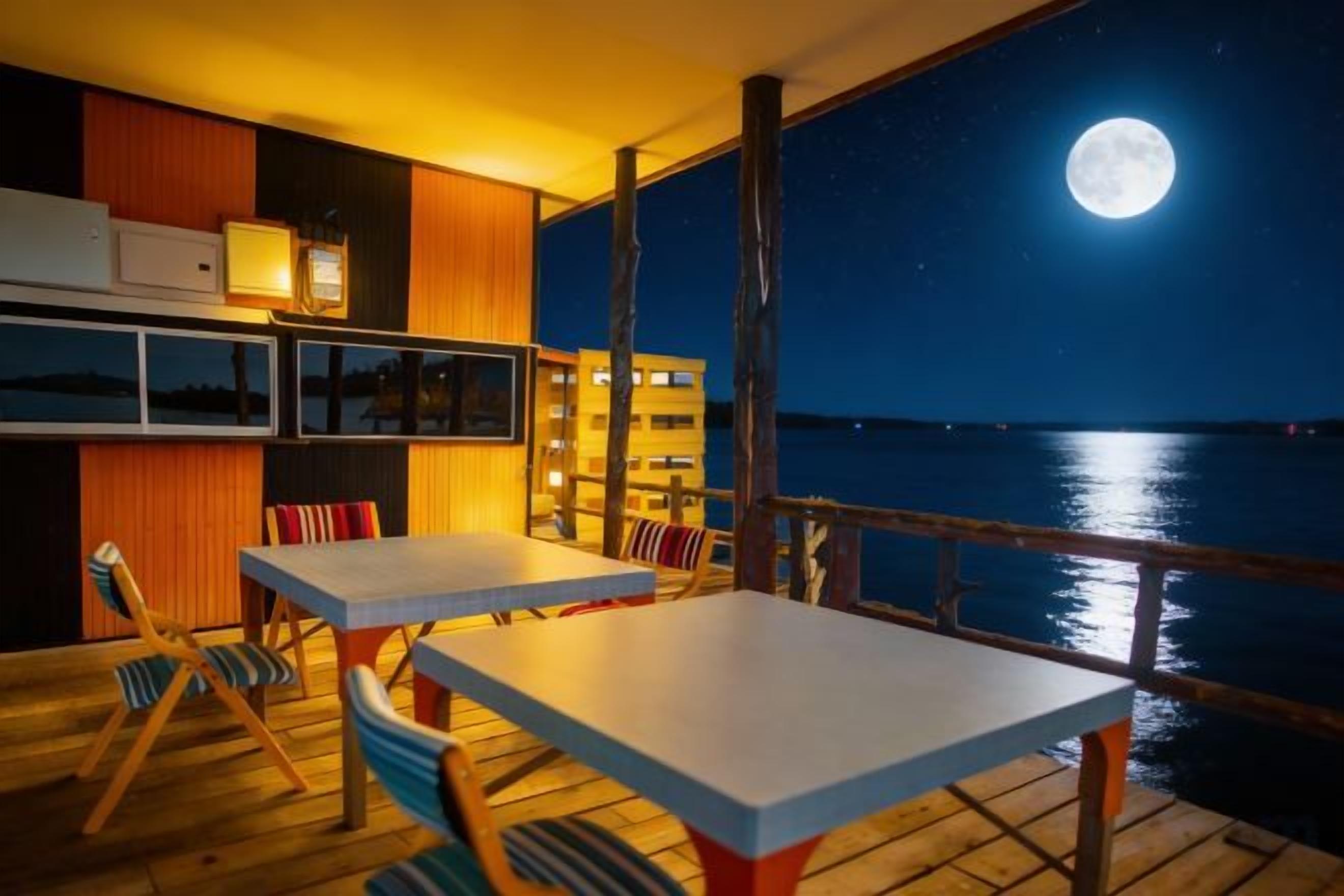 Ein Deck mit Tischen und Stühlen, das auf ein Gewässer mit Mond im Himmel blickt.