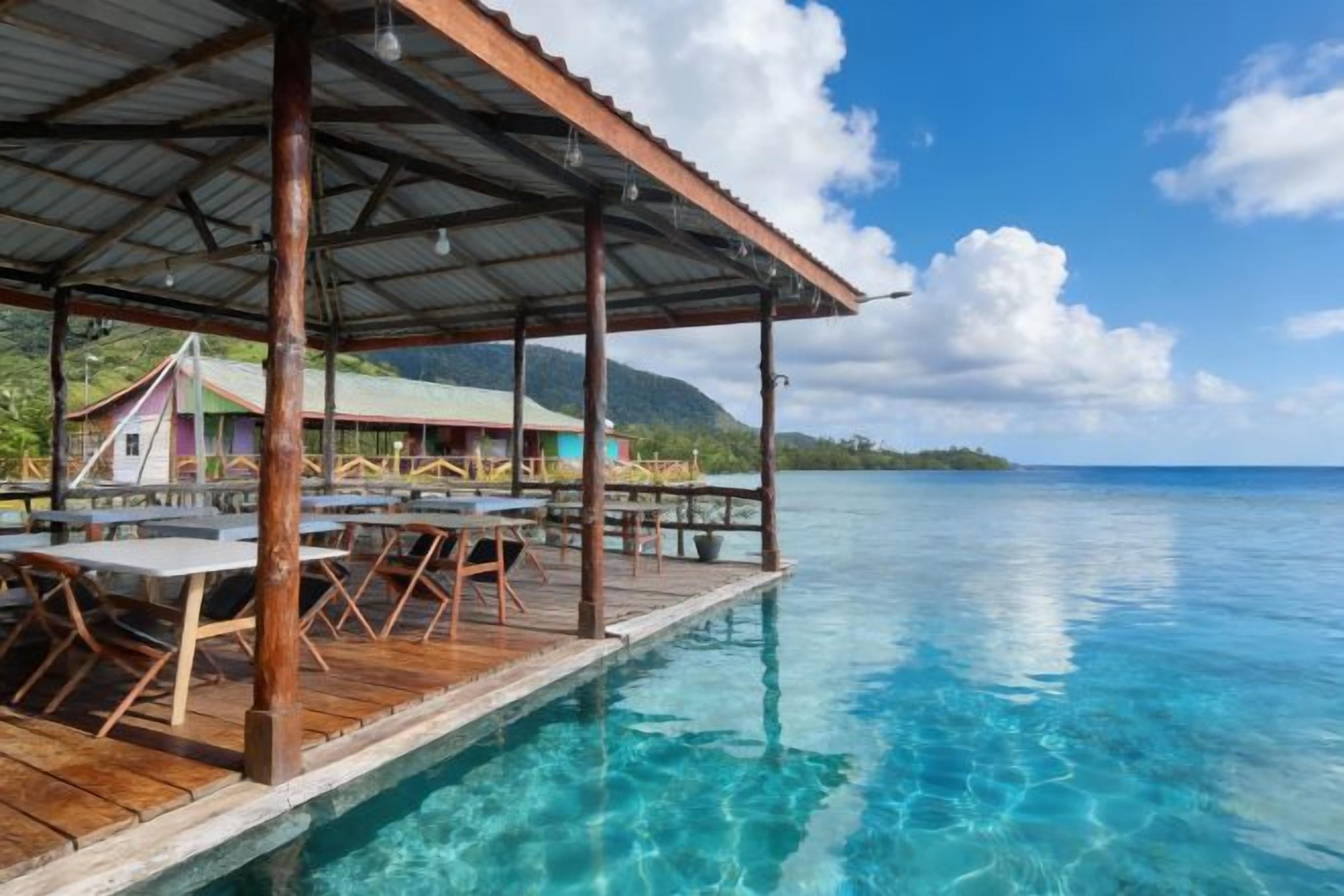 Una terrazza in legno con tavoli e sedie che si affaccia su un mare cristallino.