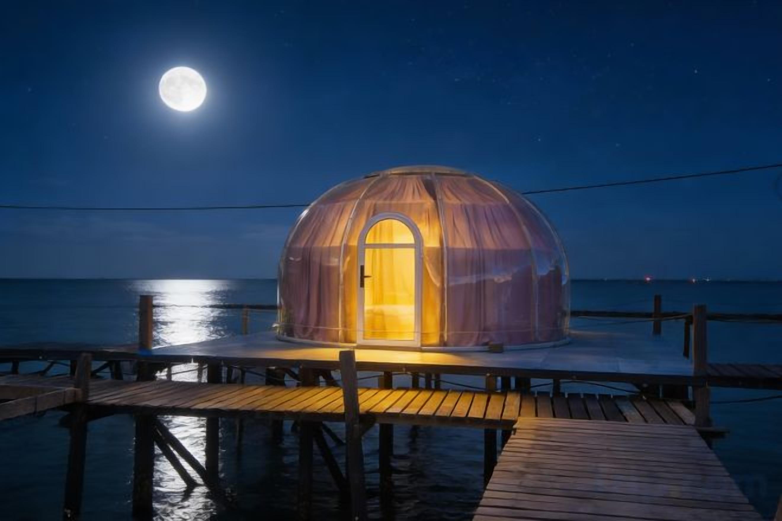 Una estructura en forma de cúpula con una puerta, ubicada sobre una terraza de madera que se extiende sobre el agua, bajo un cielo nocturno con luna visible.