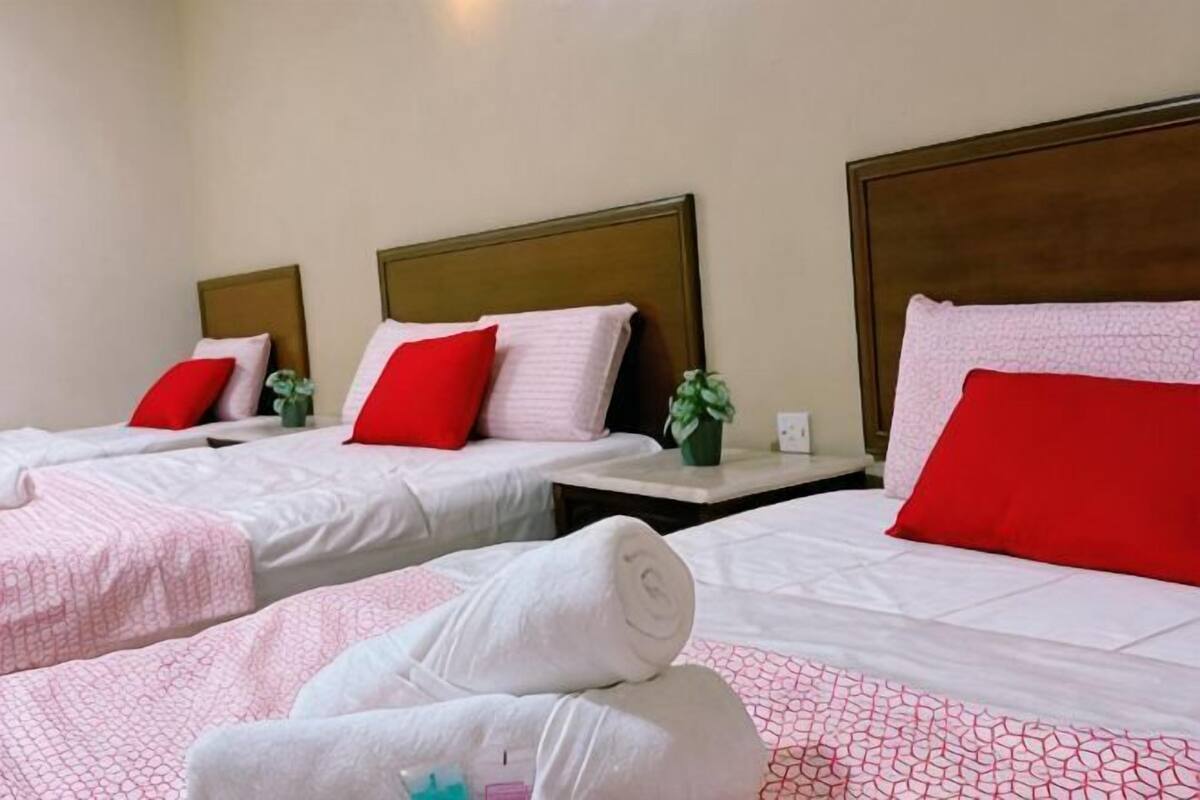 Une chambre d’hôtel avec trois lits, chacun doté d’une tête de lit en bois, de draps blancs et d’oreillers rouges. De petites tables de chevet agrémentées de plantes sont également présentes.