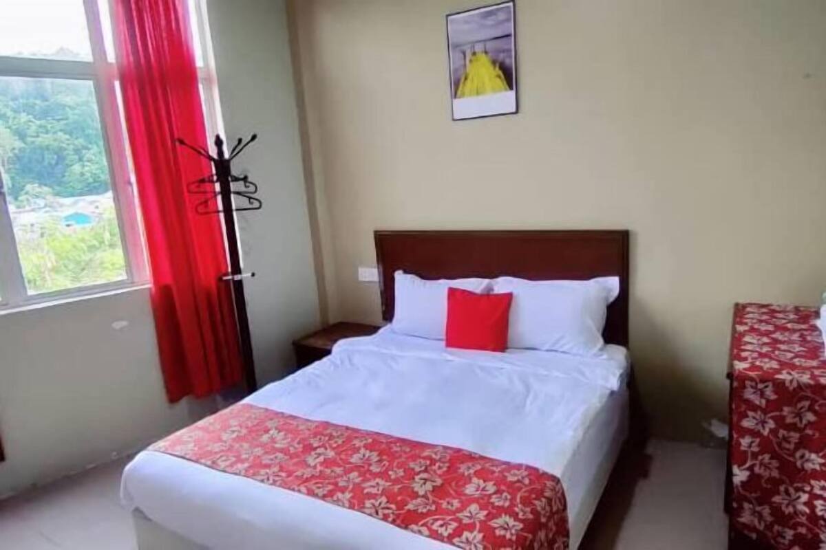 Une chambre d’hôtel avec un lit, une table de chevet, un porte-manteau, une fenêtre avec des rideaux rouges et un tableau encadré au mur.
