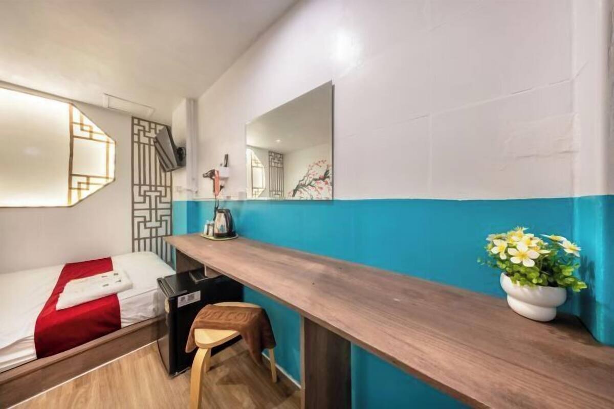 A compact hotel room with a bed, a desk, a chair, a small table with a kettle, a potted plant, and a mirror.